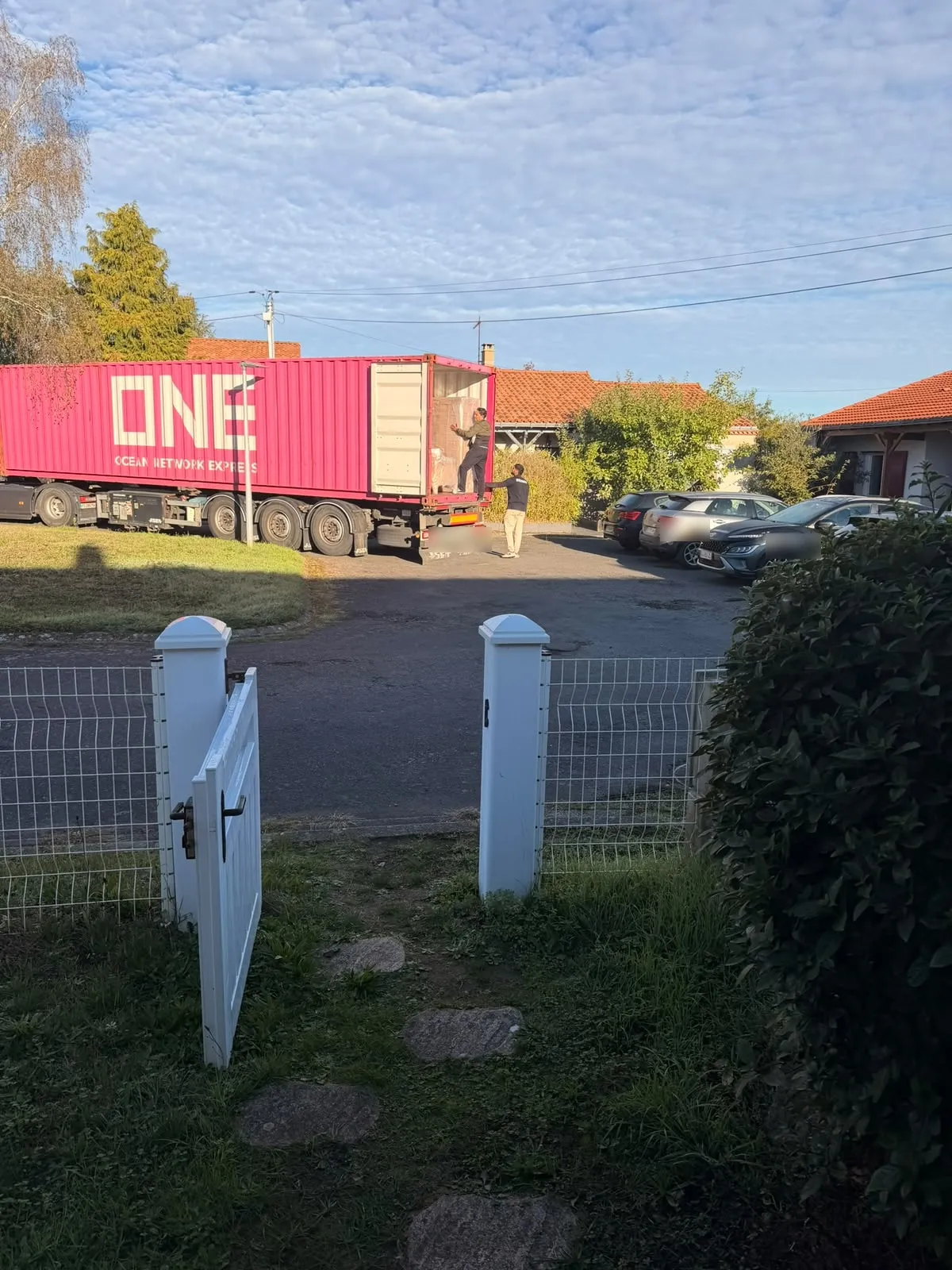 Vu depuis le portail d’un jardin, un camion porte-conteneur rose stationné dans une rue pavillonnaire, deux déménageurs à l’arrière.
