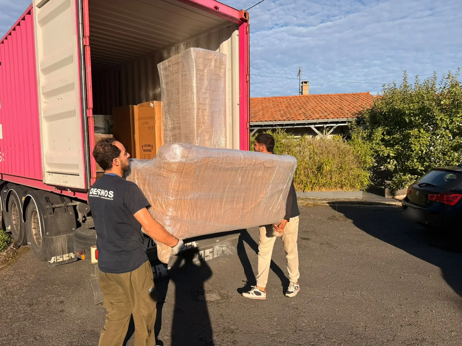 Deux déménageurs portent un canapé emballé sous film bulle vers un conteneur maritime, par un matin ensoleillé.