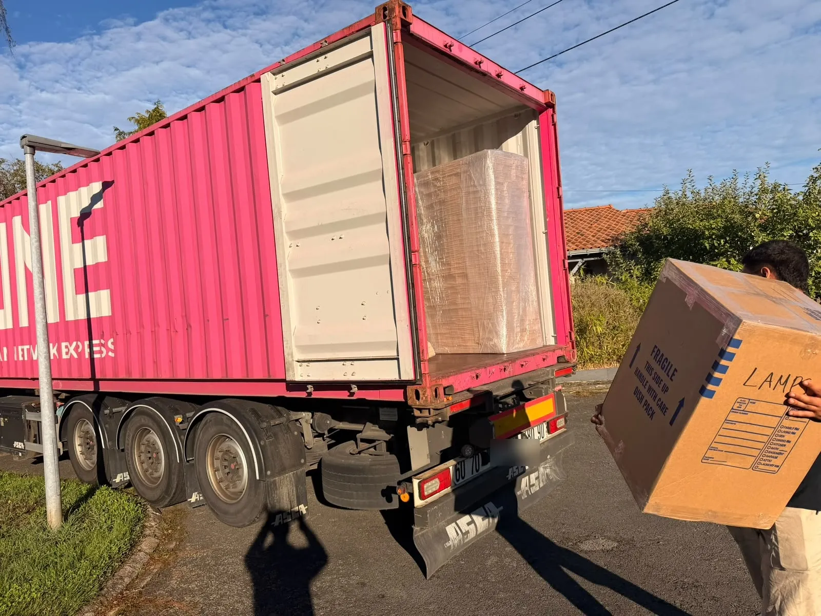 Un déménageur porte un grand carton marqué « Fragile – Lamps » devant un conteneur maritime rose dont le mobilier emballé est visible.