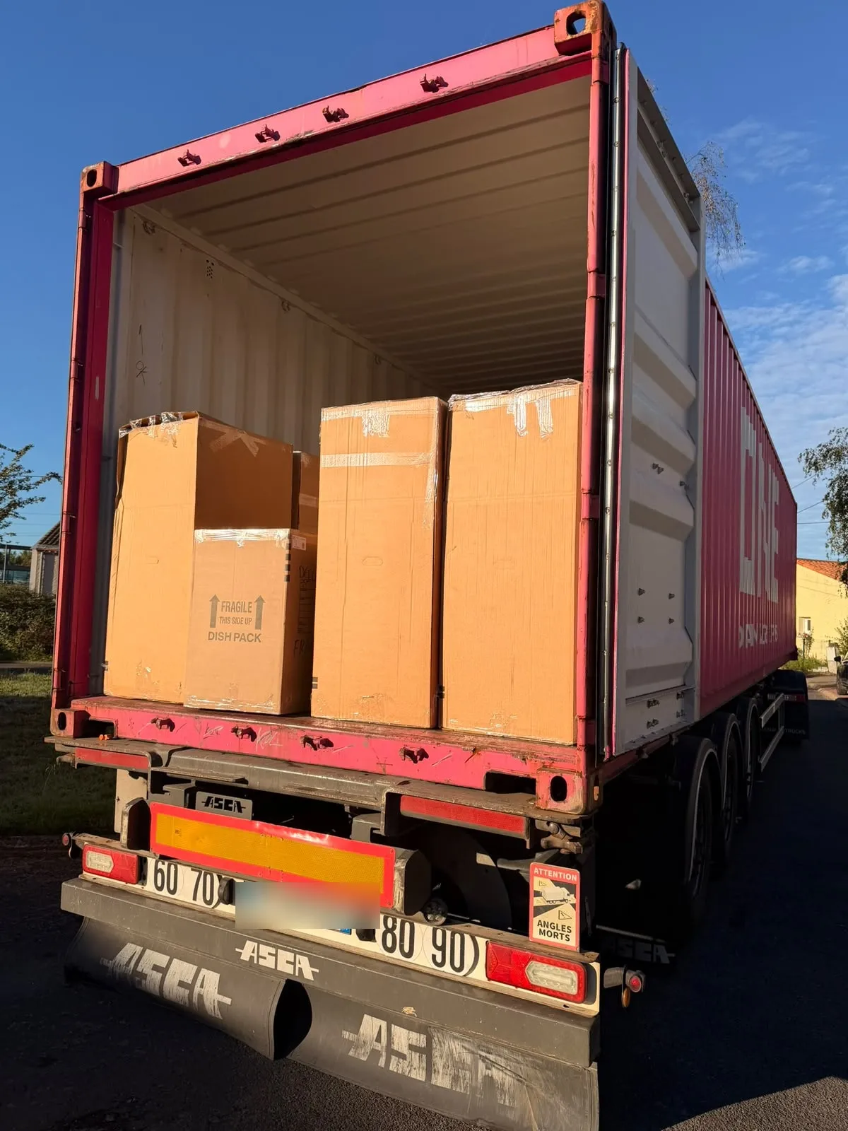 Arrière d’un conteneur maritime rose ouvert sur une remorque, dans la lumière du matin, avec de grands cartons de déménagement dont un carton « fragile » pour la vaisselle.
