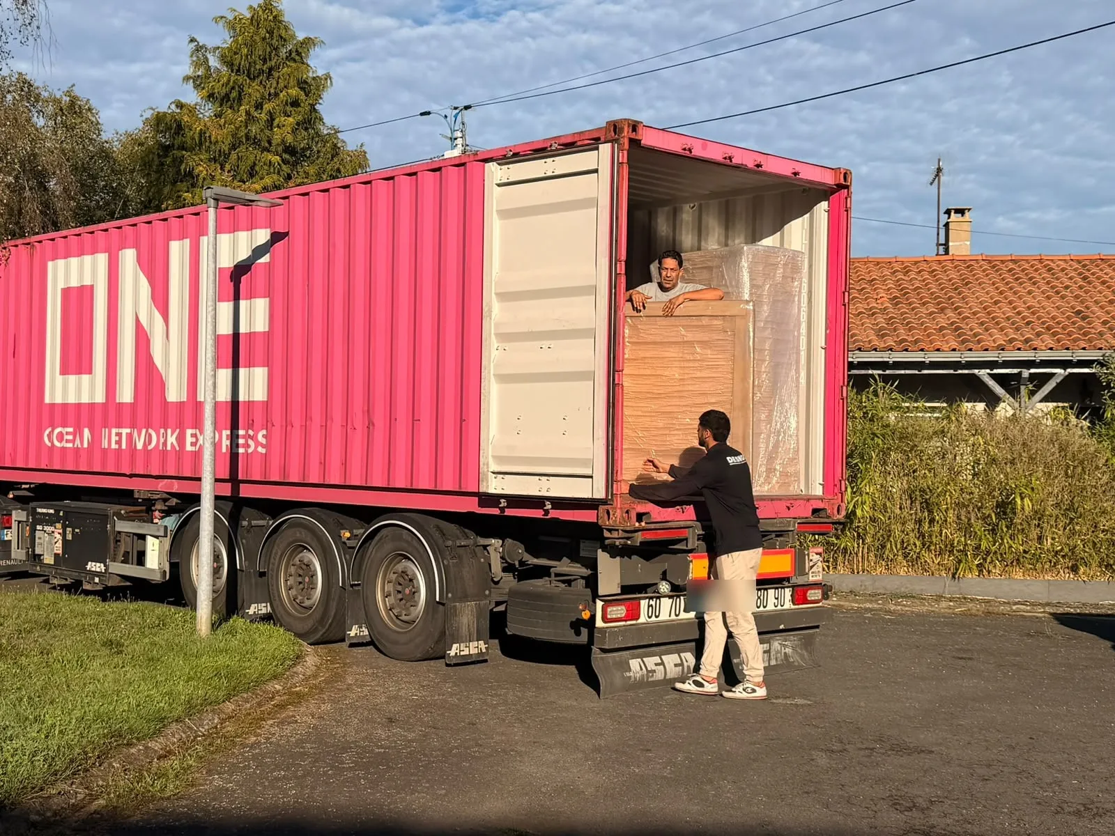 Un déménageur dans un conteneur maritime rose tend un meuble emballé à un collègue resté au sol, devant une maison aux tuiles rouges.