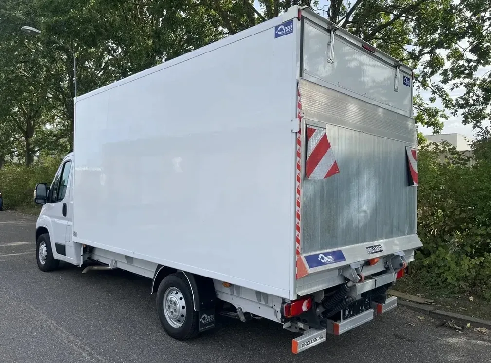 Camion fermé blanc avec hayon élévateur relevé, vu de trois quarts arrière sous les arbres.