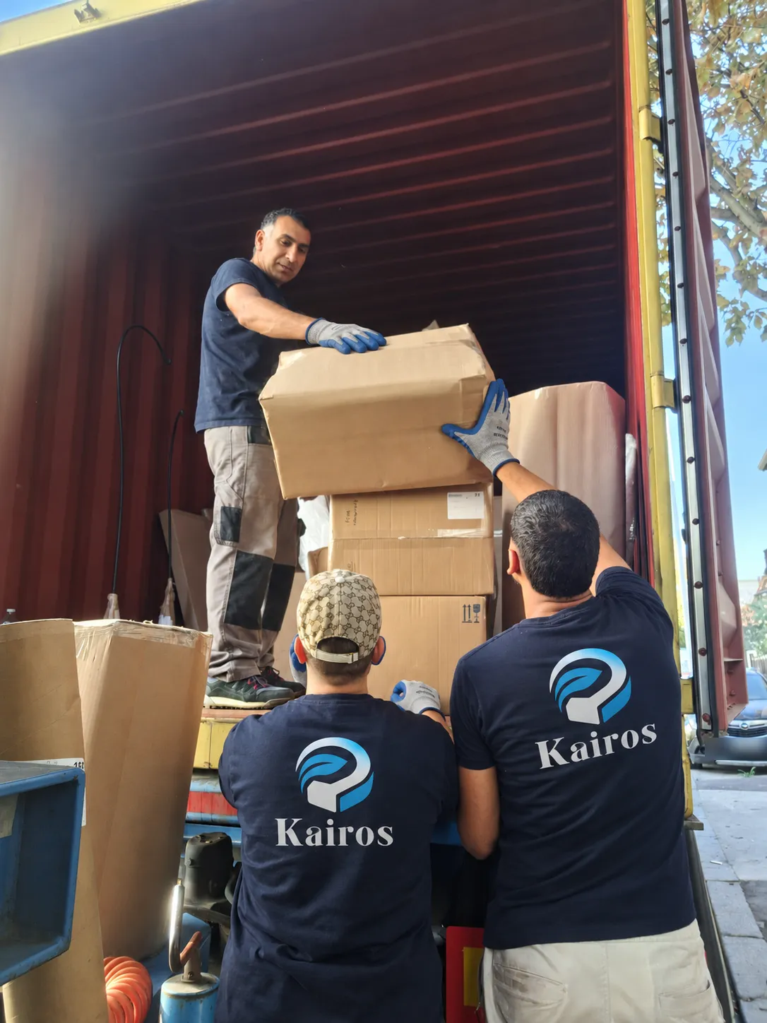 Trois membres de l’équipe KAIROS déchargent des cartons depuis un conteneur, en t-shirt bleu nuit floqué du logo.